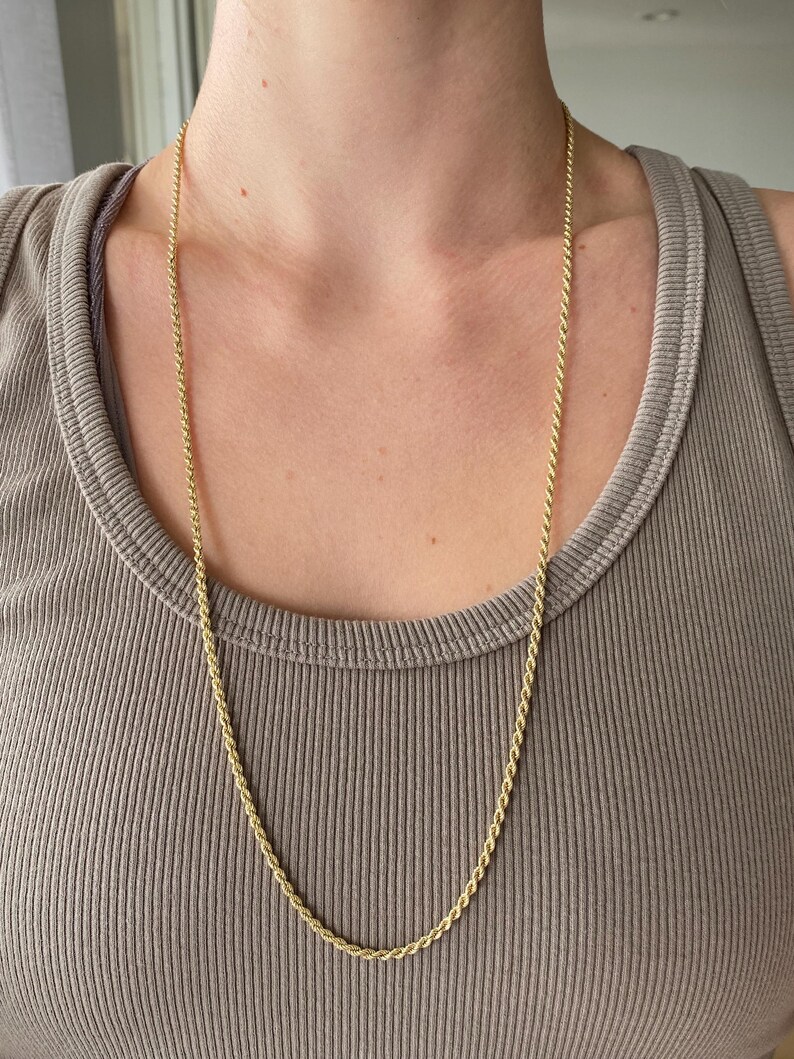 Puede incluir: Un collar largo de cadena de cuerda dorada. La cadena est&aacute; drapeada alrededor del cuello y cuelga sobre el pecho. El collar se usa sobre una camiseta sin mangas de color neutro.