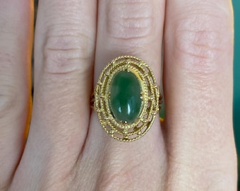 May include: A gold ring featuring an oval, deep green gemstone. The gemstone is set within a detailed, filigree-style gold setting. The ring is shown on a person's finger, highlighting the intricate design and color contrast.