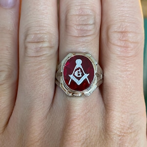 May include: A silver ring with a red center featuring a white Masonic symbol. The ring has a detailed, textured band and is set against a skin-toned background.