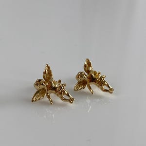 May include: A pair of gold-colored angel earrings. Each earring features a detailed depiction of a cherub with outstretched wings. The earrings are small and appear to be made of a shiny metal.