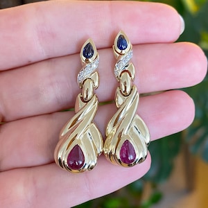 May include: A pair of gold earrings featuring a teardrop ruby at the base and a sapphire at the top. The earrings have a twisted design with a diamond-set section. The earrings are held in a hand.