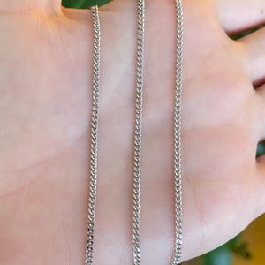 May include: Three silver curb chains are displayed against a neutral background. The chains are made of small, interlocking links, creating a textured surface. The chains are likely jewelry, suitable for wearing around the neck or wrist.