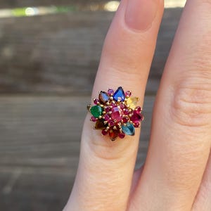 May include: A gold ring featuring a cluster of colourful gemstones, including red, green, blue, and brown. The ring is set with a variety of shapes and sizes of stones, creating a unique and eye-catching design.