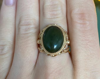 May include: A gold ring featuring an oval, dark green gemstone. The ring has an ornate, filigree design around the stone and on the band. The ring is shown on a person's finger.