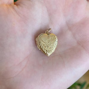 May include: A gold heart-shaped locket with intricate floral and scrollwork detailing. The locket is open, revealing a small space for a photograph or keepsake. The pendant has a small loop for a chain.
