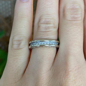 May include: A silver ring with a row of small, clear gemstones set in a channel. The ring is on a finger, showcasing the design and sparkle of the gemstones. The ring appears to be made of silver or a similar metal.