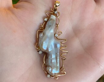 May include: A gold pendant featuring a large, irregularly shaped pearl with iridescent hues. The pearl is set within a delicate gold filigree frame, accented with small, sparkling diamonds. The pendant is designed to be worn as a necklace.
