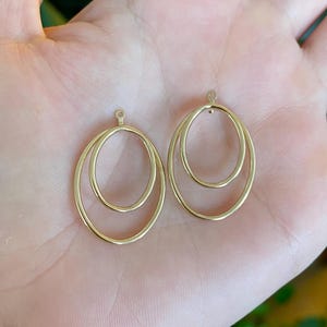May include: Two gold-colored oval-shaped earring charms. Each charm features two nested ovals, creating a layered effect. The charms have a small loop at the top for attachment. The background is a hand.