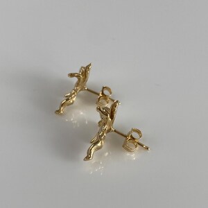 May include: A pair of gold-colored stud earrings. Each earring features a detailed, abstract design with a post and butterfly backing. The earrings are displayed on a white surface.