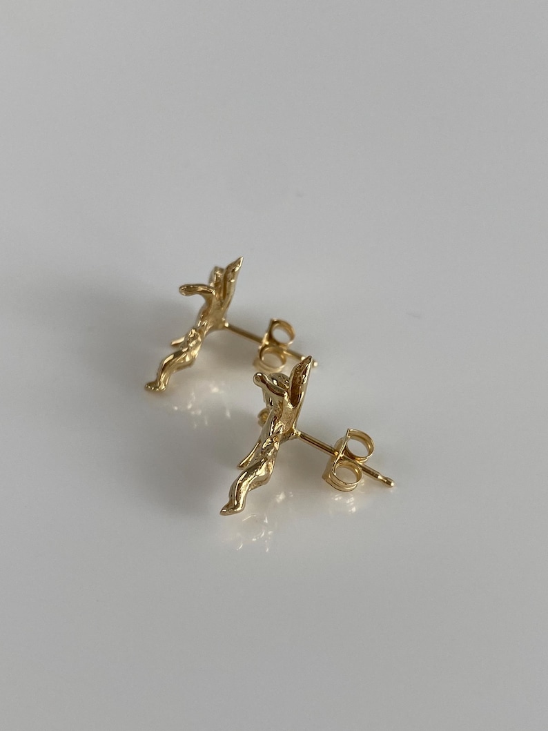 May include: A pair of gold-colored stud earrings. Each earring features a detailed, abstract design with a post and butterfly backing. The earrings are displayed on a white surface.