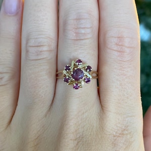 May include: A gold ring featuring a central round dark pink gemstone surrounded by six smaller round dark pink gemstones. The stones are set in a cluster arrangement with gold metal prongs and a split shank band design that encircles the finger.