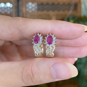 May include: A pair of gold earrings featuring oval-cut ruby gemstones surrounded by small, round diamonds. The earrings have a unique design with a cluster of diamonds around the ruby and a curved gold band.