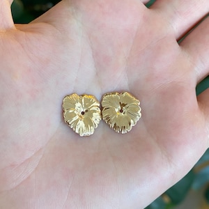 May include: Two small, gold-colored metal buttons shaped like pansy flowers. Each button features a detailed, textured petal design with a central hole for attachment. The surface of the buttons has a reflective, polished finish that catches light.