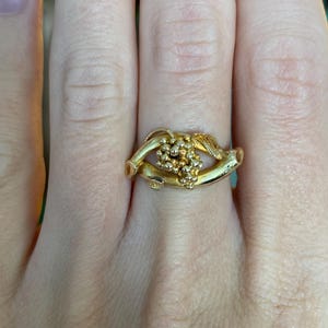 May include: A gold-colored ring featuring a cluster of small, round beads at the center. The ring's band has a unique, organic design with curved elements. The ring is worn on a finger.