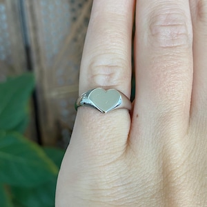 May include: A polished silver-tone signet ring featuring a smooth, flat heart-shaped face. The band is solid and tapers slightly toward the back, showcasing a clean, reflective metallic surface that sits flush against the finger.