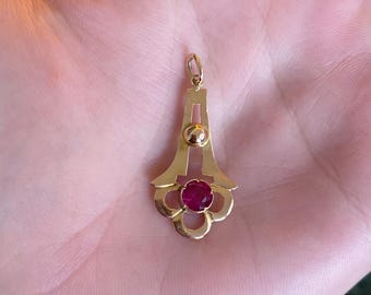 May include: A gold pendant with a ruby gemstone. The pendant has a floral design with a central red gemstone and a small gold ball. The pendant is suspended from a small loop.