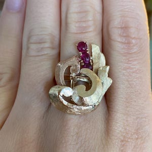 May include: A gold-toned ring featuring a textured, swirling design. The ring is adorned with three oval-shaped, deep red gemstones. A small, clear gemstone is also visible.