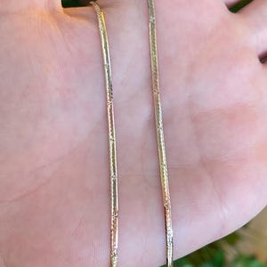May include: A gold-toned herringbone chain necklace featuring a flat, flexible design with a subtle, repeating etched star pattern along the surface of the metal links. The chain has a polished, reflective finish that catches the light.