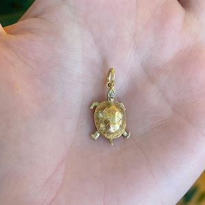 May include: A gold-colored turtle pendant with a small diamond accent. The pendant features a detailed turtle shell design, four tiny legs, and a loop for a chain. The turtle's head has a small diamond. The pendant is likely made of gold.