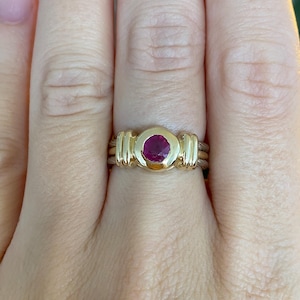 Peut inclure: Une bague en or ornée d'une pierre précieuse ronde d'un rouge rosé profond, montée dans un serti clos. L'anneau est composé de plusieurs fils d'or parallèles qui convergent vers une section en or lisse et solide retenant la pierre.
