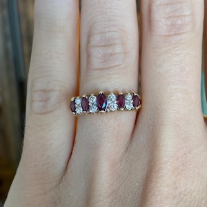 Peut inclure: Une bague en or ornée d'alternances de pierres rouges ovales et de petites pierres claires rondes. La bague est présentée sur une main avec une manucure aux tons neutres, mettant en valeur le design et le contraste de couleurs.