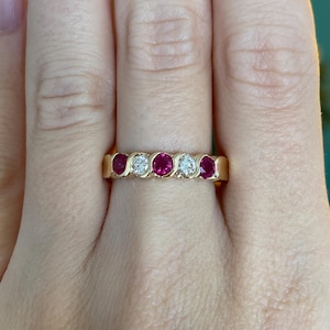 May include: A gold band ring featuring alternating round-cut rubies and diamonds. The ring has a classic design with the gemstones set in a row. The rubies are a deep red color, and the diamonds sparkle brightly. The ring is shown on a person's finger.