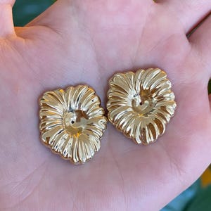 May include: Two gold-toned metal buttons with a raised, radiating petal design. Each button features a smooth, reflective surface with a central indentation and a small hole in the middle, creating a flower-like appearance with defined ridges.