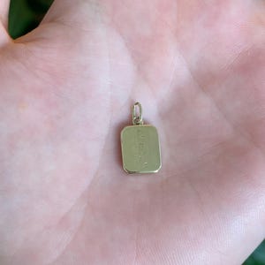 May include: A small rectangular gold pendant featuring a vertical striped texture on its front surface. The pendant has rounded corners and includes a small circular jump ring at the top for attachment to a chain or necklace.