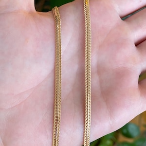 May include: A gold-toned herringbone chain necklace featuring a flat, woven link design with a polished, reflective surface. The chain displays a consistent, tight chevron pattern throughout its length, creating a smooth and flexible metallic texture.