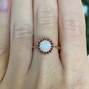 May include: A gold ring featuring a central round white pearl surrounded by a halo of small, deep pink rubies. The setting is circular, and the band is a thin, polished gold metal that tapers toward the back of the finger.