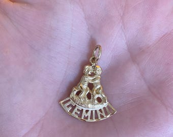May include: A gold-colored Gemini pendant. The pendant features two figures embracing, with the word "GEMINI" in a cutout design at the bottom. The pendant is held in a hand.