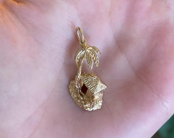 May include: A gold-colored pendant featuring a tropical island scene. The pendant depicts a small house with a palm tree, and a small red stone. The pendant is likely made of gold and is designed to be worn as jewelry.