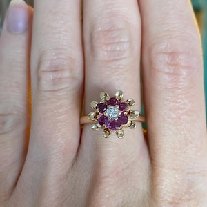 May include: A gold ring with a flower-like design. The centre of the ring has a small, round, clear diamond, encircled by a cluster of deep pink gemstones. The ring's band is a simple, polished gold.