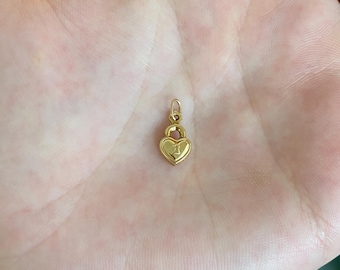 May include: A small gold-toned heart-shaped padlock charm featuring a central keyhole cutout. The charm has a smooth, polished finish and includes a small circular jump ring at the top for attachment to a chain or bracelet, presented in a palm.