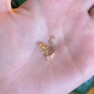 May include: A small gold charm in the shape of a spinning wheel, featuring a circular wheel component attached to a frame with four legs and a small jump ring at the top for attachment to a chain or bracelet.