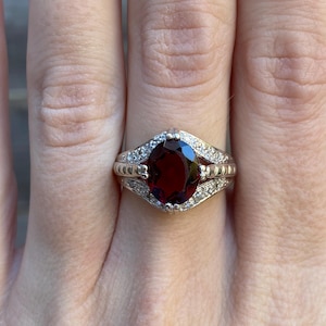 May include: A silver ring featuring a large oval red gemstone, surrounded by smaller clear gemstones.