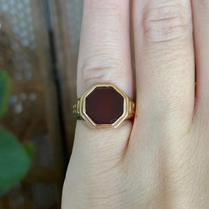 May include: A gold signet ring featuring an octagonal-shaped dark red carnelian stone set in the centre. The band of the ring displays a decorative geometric pattern on the sides, providing a structured and polished appearance to the jewellery piece.