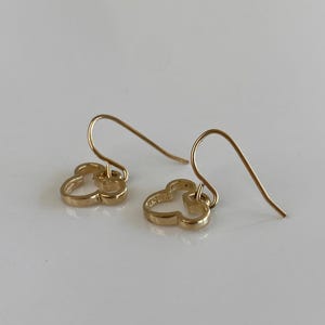 May include: A pair of gold-colored earrings. Each earring features a clover-shaped design with a hook closure. The earrings are displayed on a white surface, with the light reflecting off the gold metal.