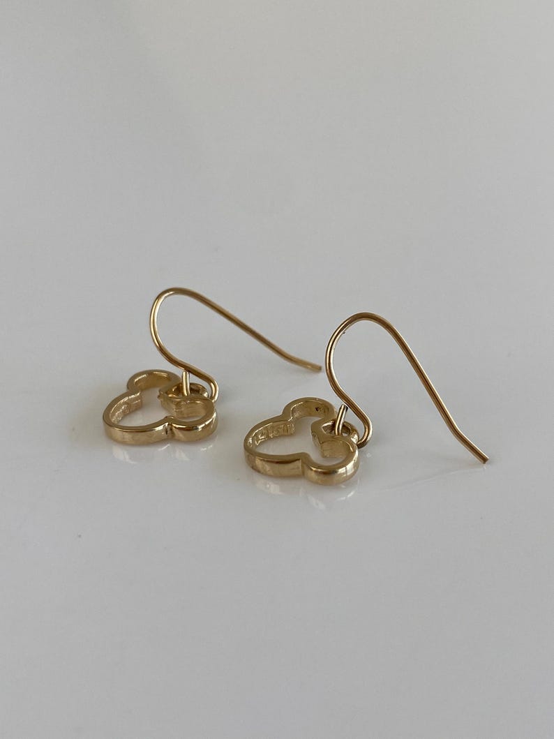 May include: A pair of gold-colored earrings. Each earring features a clover-shaped design with a hook closure. The earrings are displayed on a white surface, with the light reflecting off the gold metal.