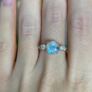May include: A gold ring featuring a large, iridescent, round gemstone as the centerpiece. The ring is flanked by smaller, clear gemstones on each side. The ring is set on a hand, showcasing the design and sparkle of the jewelry.
