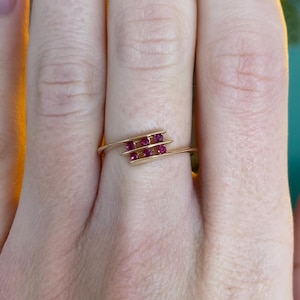 May include: A gold ring with a distinctive design. The ring has two parallel bars, each set with three small, bright red gemstones. The ring is worn on a finger, highlighting its delicate and elegant style.