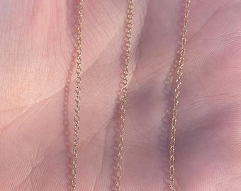 May include: A gold chain necklace with a delicate, thin chain link design. The chain is shown laid out on a person's hand.
