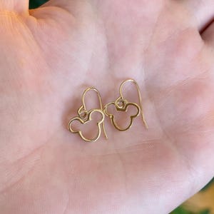 May include: A pair of gold-colored, open-design earrings in the shape of a mouse head. The earrings are held in a person's hand, with the hook of the earring visible. The earrings are a simple, elegant design.