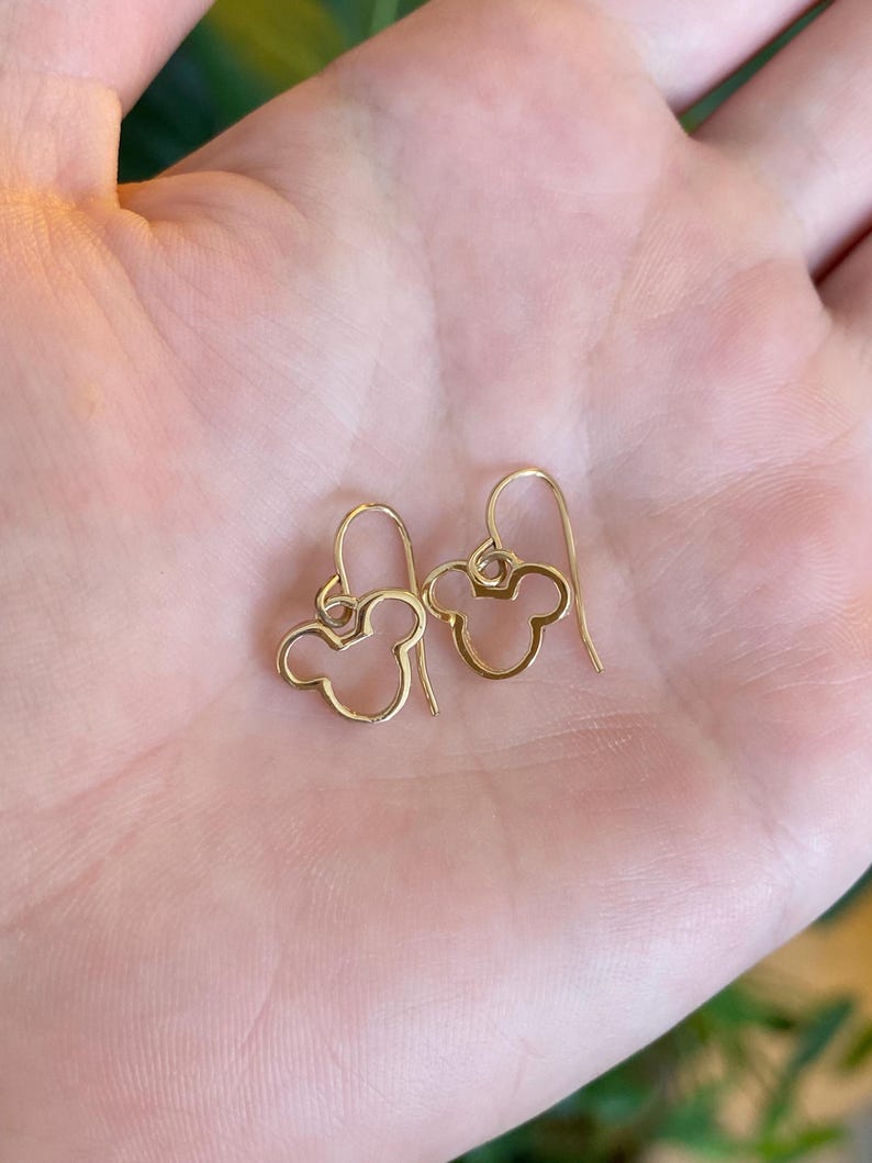 May include: A pair of gold-colored, open-design earrings in the shape of a mouse head. The earrings are held in a person's hand, with the hook of the earring visible. The earrings are a simple, elegant design.
