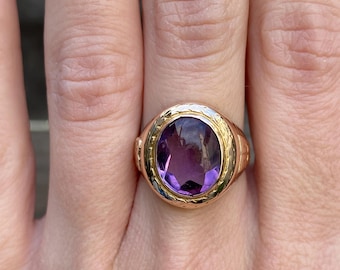 May include: A gold ring with a large oval amethyst gemstone set in a textured bezel. The ring has a detailed band.