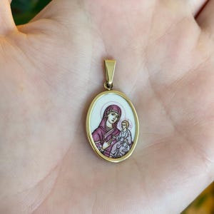 May include: Gold-tone oval pendant featuring a religious icon of a woman and child. The pendant has a white background with a gold border and a small gold bail. The woman is wearing a purple head covering and holding the child.
