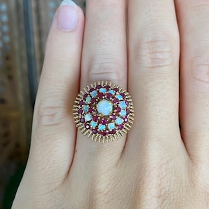 May include: A gold ring featuring a central round opal surrounded by concentric circles of small round rubies and opals. The setting has a textured, rope-like gold border, creating a circular cluster design that sits prominently on the finger.