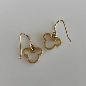 May include: A pair of gold-colored dangle earrings. Each earring features a cutout design of a three-circle silhouette, resembling a famous cartoon character's head. The earrings have a simple hook closure. The background is a plain white surface.
