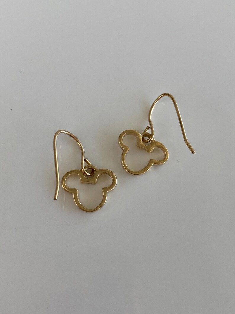May include: A pair of gold-colored dangle earrings. Each earring features a cutout design of a three-circle silhouette, resembling a famous cartoon character's head. The earrings have a simple hook closure. The background is a plain white surface.