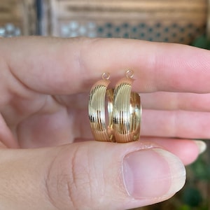 May include: A pair of gold-toned hoop earrings featuring a polished finish with vertical ribbed texturing along the outer surface. Each earring has a small circular loop at the top for attachment, showcasing a curved, semi-circular design.